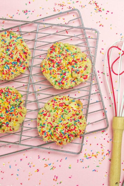 sugar cookies with lots of sprinkles sit on baking racks with a heart-shaped whisk next to the rack; there is a pink background.