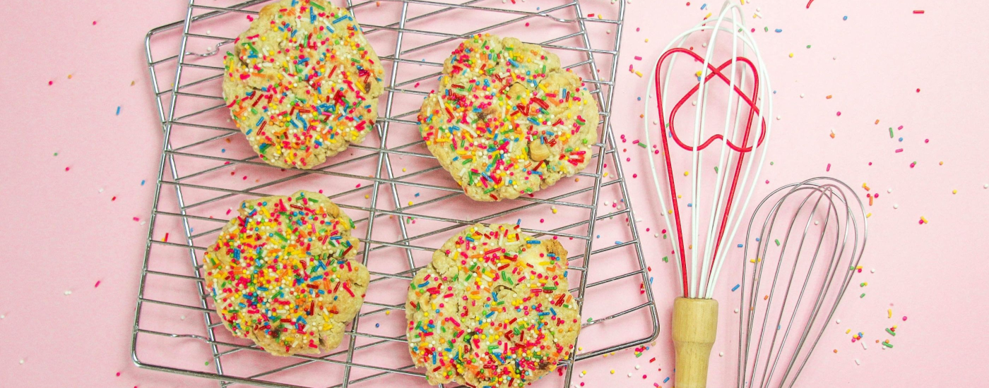 sugar cookies with lots of sprinkles sit on baking racks with a heart-shaped whisk next to the rack; there is a pink background.