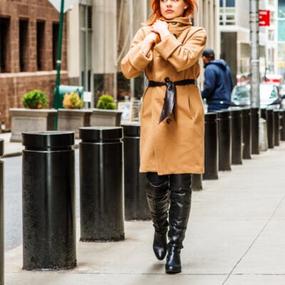 professional woman walks along city street; she is wearing black patent leather knee-high boots and a camel-colored wool overcoat