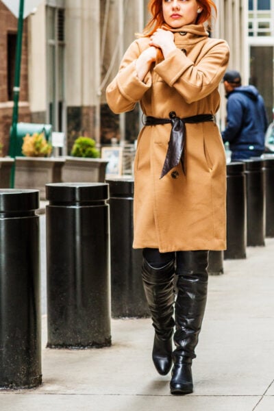 professional woman walks along city street; she is wearing black patent leather knee-high boots and a camel-colored wool overcoat