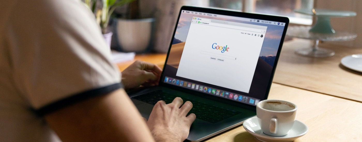 person works on laptop computer with browser open to Google; they sit at a desk with a white cup of coffee to their right and a green plant in the distance