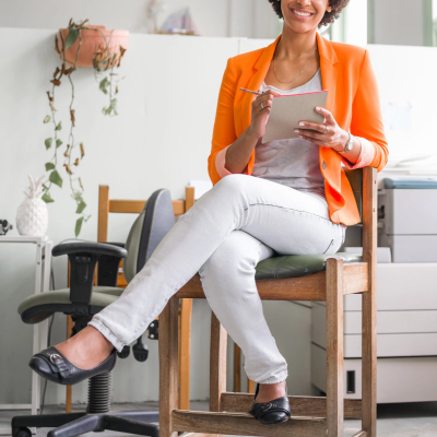 Black businesswoman wears comfortable ballet flats, white pants, and a bright orange blazer; she is working in a business casual office