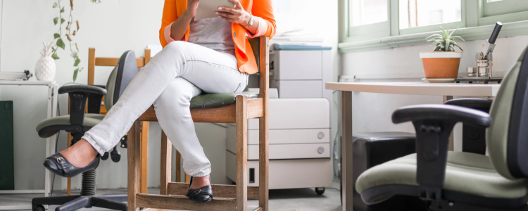 Black businesswoman wears comfortable ballet flats, white pants, and a bright orange blazer; she is working in a business casual office