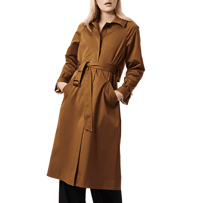 woman wears brown single-breasted trench coat; it is belted and she has her hands in the pockets