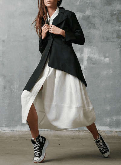 woman wears black asymmetric blazer with a white midi dress and Converse; the model has her legs in a weird position like she's going to start running.