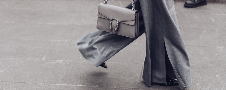 woman in gray suit with very wide leg pants walks along city street; she is holding a gray shoulder bag