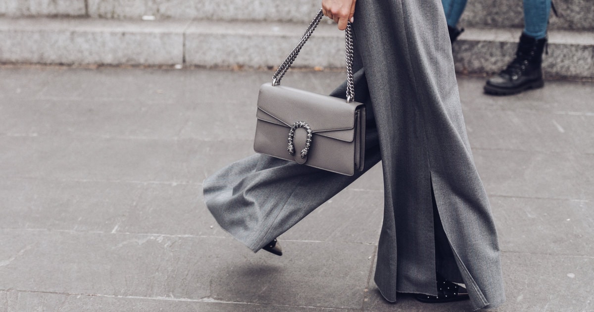 woman in gray suit with very wide leg pants walks along city street; she is holding a gray shoulder bag