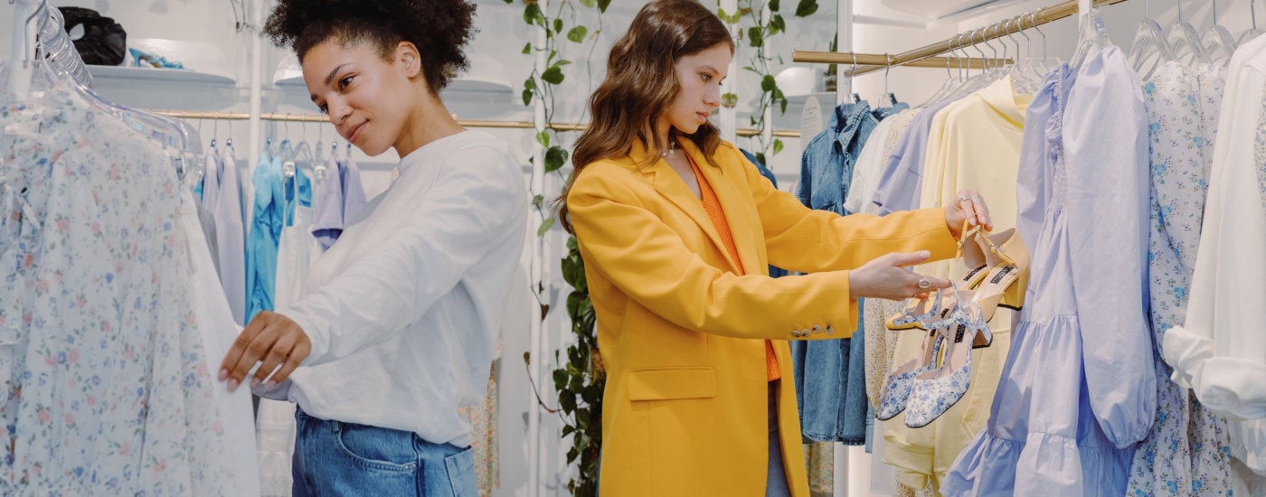 two women shop for clothes to step up their work wardrobes; one woman is black and wearing a whiteish top and jeans, the other woman is white and wearing a yellow blazer and orange shirt