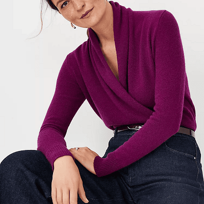 woman wears a dark purple shawl collar faux wrap sweater with blue jeans; she is seated and hunching forward