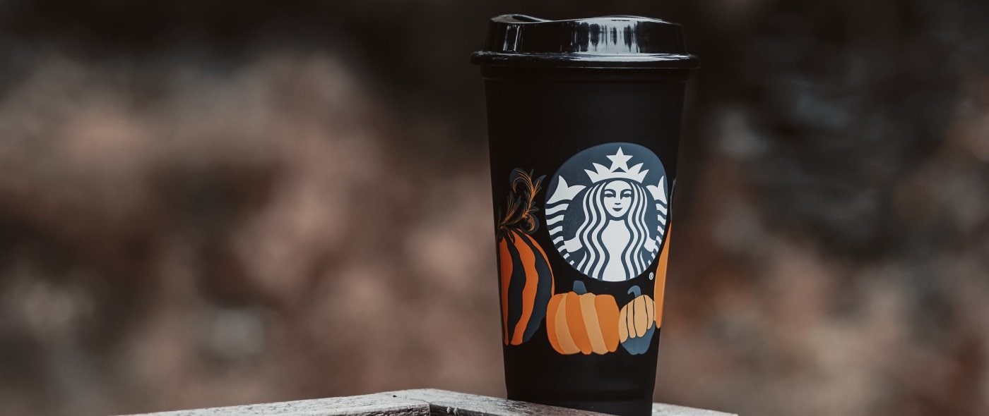 black Starbucks cup with pumpkins on it sits on top of a granite counter; there is a very blurry brownish background. 