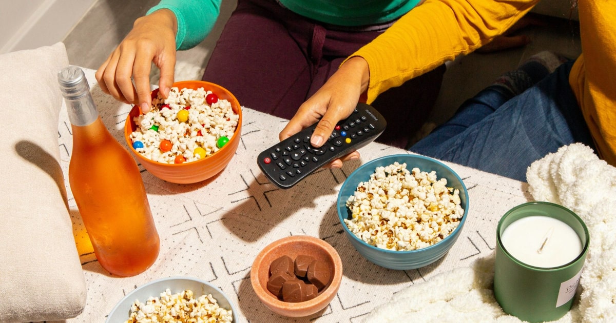 A coffee table with snacks, a bottle of wine, and a candle; two people's hands are visible, one holding a TV remote 