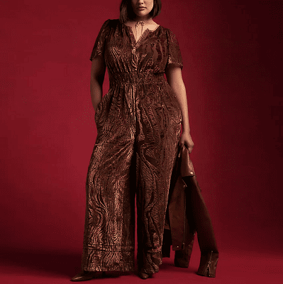woman wears dark brown velvet burnout jumpsuit; she is standing in front of a dark red background