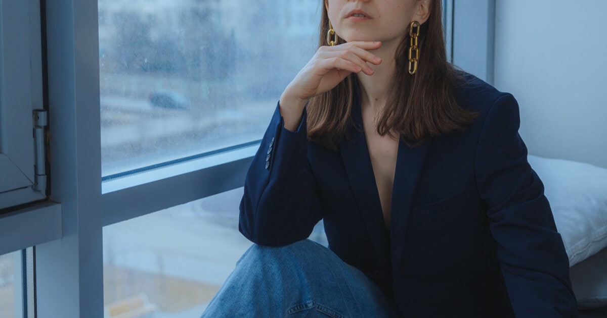 woman has adapted her conservative workwear to fit a business casual office; she wears a blazer with jeans and sits in front of a windowseat; she has dangly gold earrings on