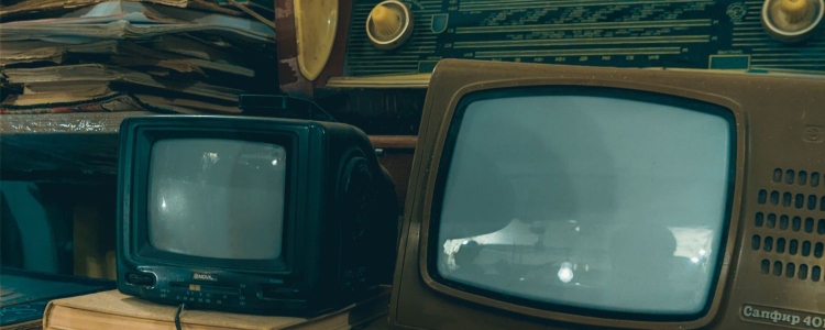 old televisions are piled on top of books; an old radio sits on the shelf above, next to magazines