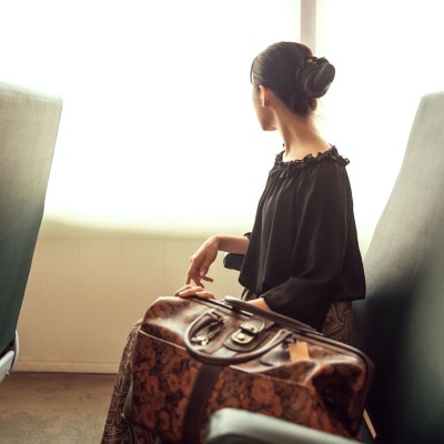 woman with black hair looks out a window; she has a weekender bag next to her and has her hair in a bun