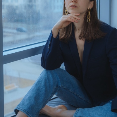 woman has adapted her conservative workwear to fit a business casual office; she wears a blazer with jeans and sits in front of a windowseat; she has dangly gold earrings on