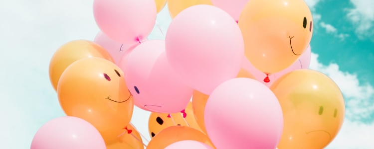 pink and orange balloons with happy faces float against a cloudy blue sky