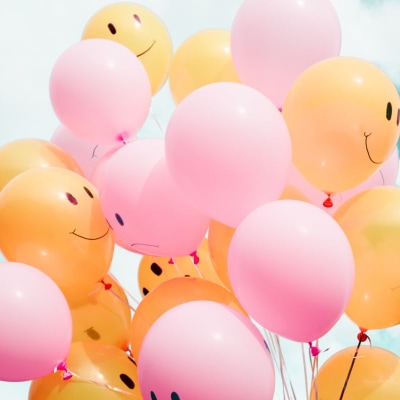 pink and orange balloons with happy faces float against a cloudy blue sky