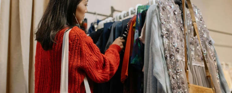 woman looks through a thrift store rack; she is trying to make her workwear more eco-friendly. She is wearing a red sweater and has a cream canvas bag slung over her shoulder.