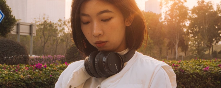 young professional woman wears headphones around her neck; she is sitting outside in a garden with a city in the distance