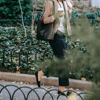 Black woman walks outside on her way to work; she is wearing what could be some of the best work pants for women in 2024! She's carrying a tote bag and drinking a coffee while she walks behind a short circular fence