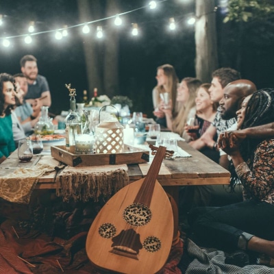 a group of friends sit around a table at night; there are string lights in the background and a large string instrument in the foreground