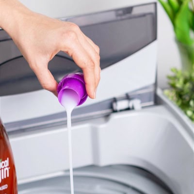 woman pours capful of laundry detergent into top-loading washer; the laundry detergent cap is purple, there is a reddish bottle of detergent on the left, and some green plants on the right