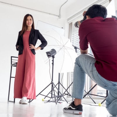woman poses for a corporate headshot or LinkedIn photo; she is wearing a pink jumpsuit with a blazer and being photographed in a white space by a kneeling photographer wearing a red shirt