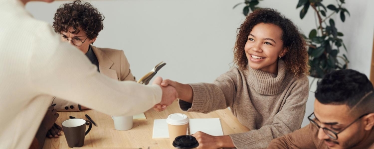 professional woman shakes someone's hand; she's read some great networking books for professional women! The Black model is wearing a brownish turtleneck sweater; she is seated next to a man wearing glasses, and the person wearing white shaking her hand is standing and cutting off the view of a curly-haired White woman wearing a beige blazer and black turtleneck.