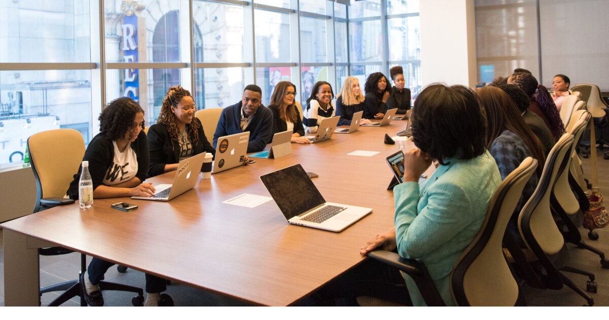 A diverse group of people dressed in business casual, at a long table having a meeting. They're in a large meeting room and have laptops in front of them.