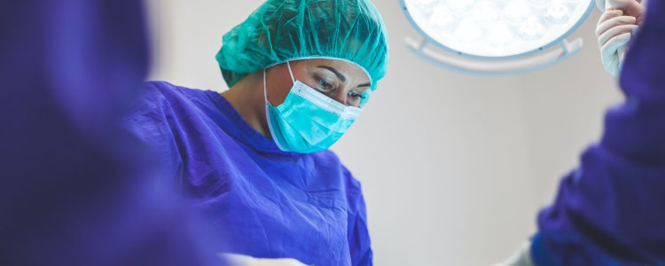 female surgeon operates on a patient; she wears a dark blue surgical gown and a lighter blue mask and hair covering