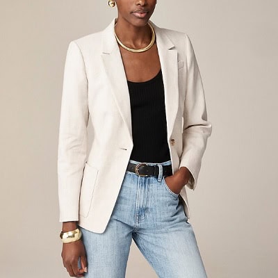 A woman wearing a cream blazer with black inner top and denim pants with belt and gold necklace, earrings and bracelet