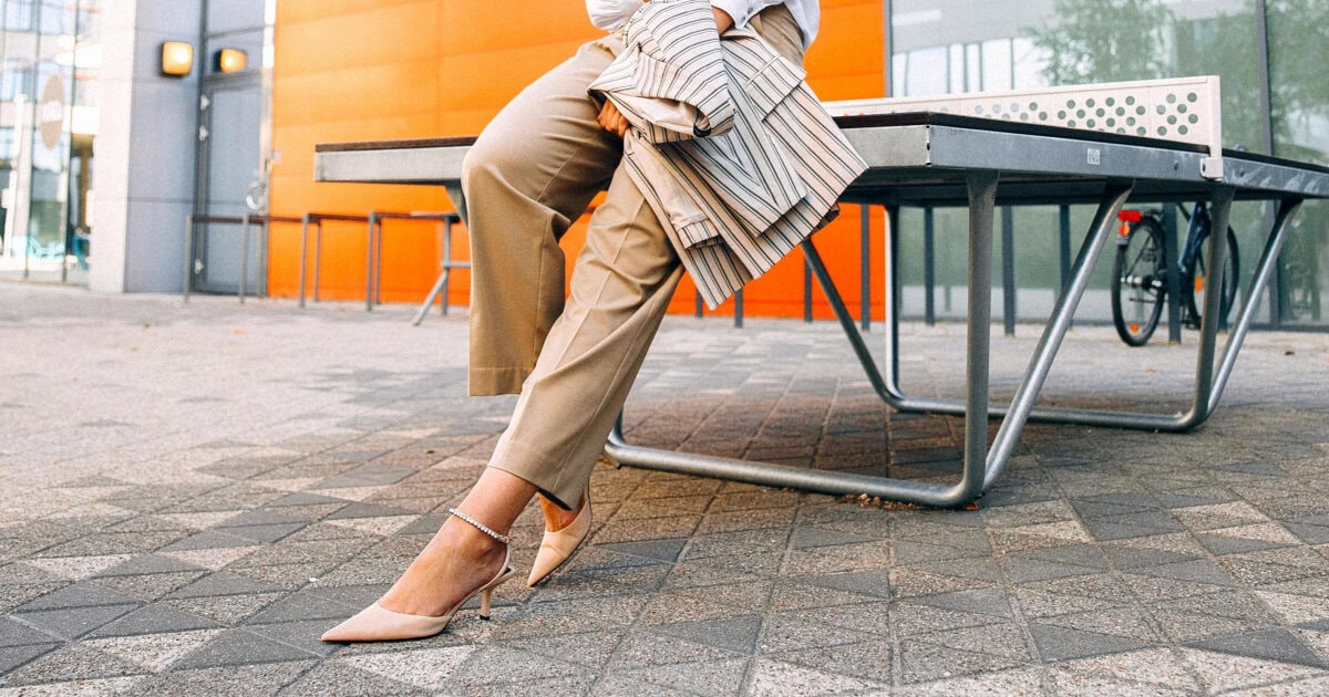professional young woman wears stylish summer work outfit in beige pants, nude heels, white blouse and striped blazer; she is leaning against a table outside on cobblestones; in the background is an orange wall