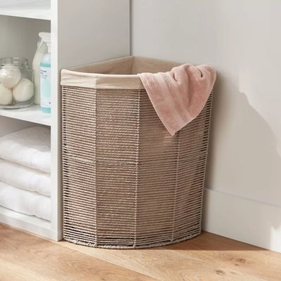 A tan woven hamper in a room next to a white shelving unit with towels and cleaning products. There is a light pink towel draped over the hamper.