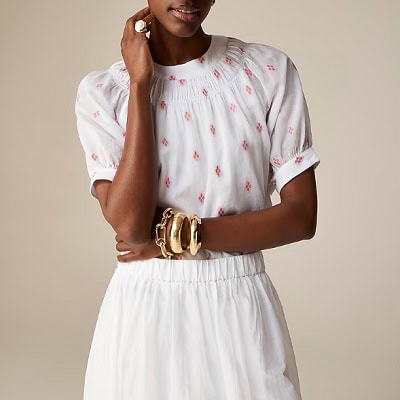 A woman wearing a white top with red print details and white skirt with gold bracelets