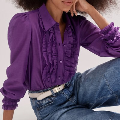 A woman wearing a purple ruffle blouse, blue jeans, and a white belt