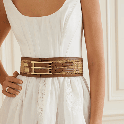 woman wears wide belt with two skinny straps; the belt is a mix of brown leather and woven raffia. The woman is wearing a white dress with corset-style seaming and standing in front of a creamy white wall with decorative wall trim