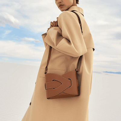 woman models a tan leather bag with an intricate knot detail across the time; she appears to be wearing a yellowish beige overcoat while walking across a white sand beach