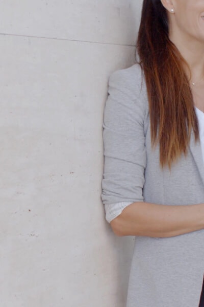 professional young woman wears summer business casual and leans against wall outside her office, talking on cell phone; she has a ponytail in her hair and a gray blazer over a white blouse