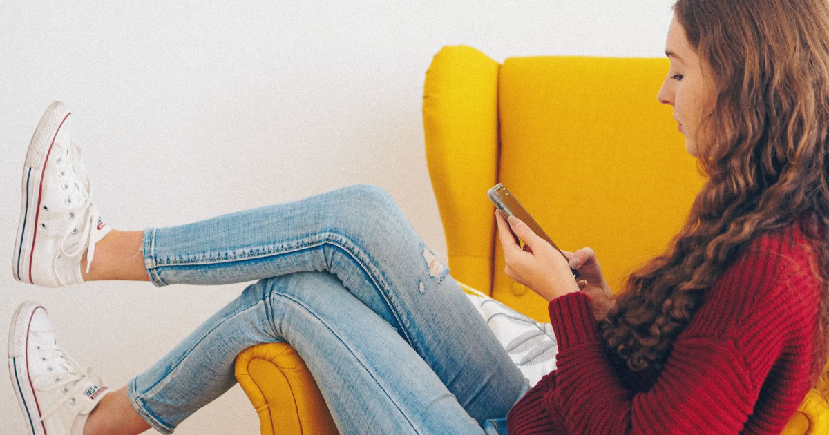 A woman in a red sweater and jeans sitting on a red easy chair while using her phone