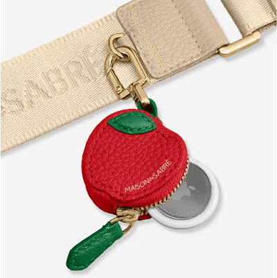 red leather charm with zipper to hold an Apple AirTag; the zipper is green and there is another green leather detail designed to look like an apple. Tiny letters on the charm (and beige strap in background) read "Maison de Sabre."