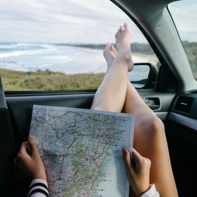 young woman reads a map while she relaxes in her car; she has her feet out the window and there is a beautiful view of water and rolling hills in the distance