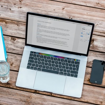 laptop on blonde wood table; there isa cup of water and a notebook to the left and a cell phone to the right