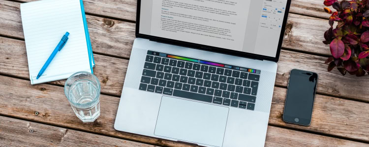 laptop on blonde wood table; there isa  cup of water and a notebook to the left and a cell phone to the right