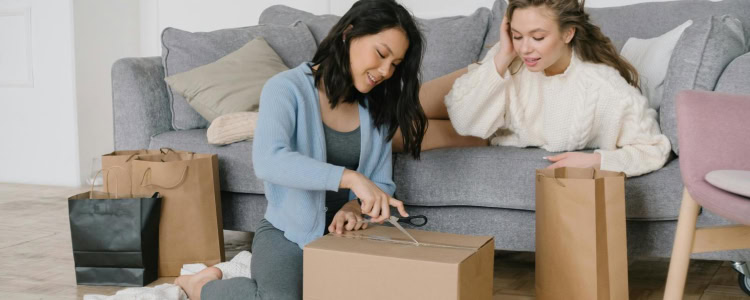 two 20-something women relax in a living room, opening packages with excitement