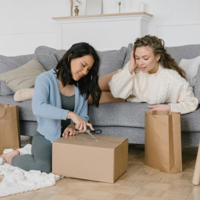 two 20-something women relax in a living room, opening packages with excitement