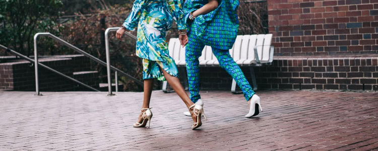 Two women walking outdoors next to each other on a brick walkway; they are wearing blue, white, and green outfits and both wearing white heels