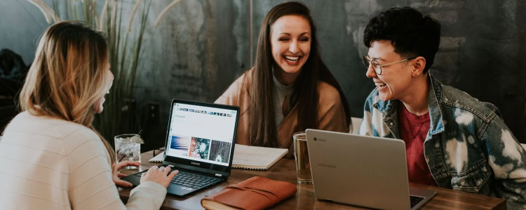 three professional young women laugh and talk at a shared table in the office