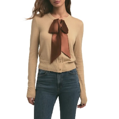 A woman wearing a tan long sleeve cardigan with brown silk bow and blue jeans