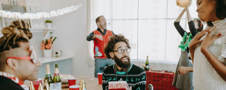 coworkers exchange white elephant gifts; in the foreground of the photo a man with a beard and glasses holds a present with red and white wrapping while a professional woman stands nearby with her arms pressed to her chest as if to say "for me?"; she is wearing a white short-sleeved sweater and beige trousers. Off to the side another coworker wears silly red holiday sunglasses. In the background a woman is throwing a balloon in an air while a man wears a red silly holiday vest while standing in front of a window.